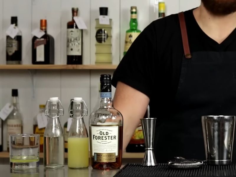 Na drvenom stolu prikazani Sastojci za Whiskey Sour i barmen u pozadini