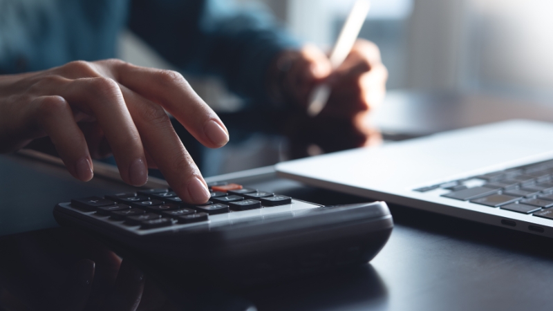 Osoba koristi digitron pored laptopa dok radi finansijske proračune, što simbolično prikazuje temu kako izabrati knjigovođu u Beogradu