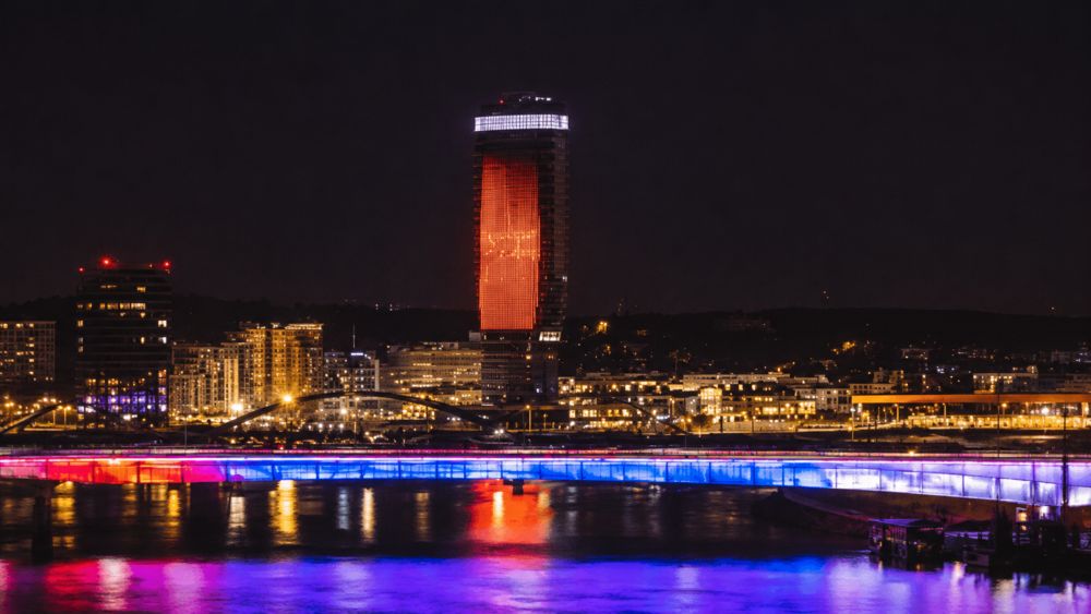 Noćni pogled na Beograd sa osvetljenim mostom preko Save i modernim zgradama u pozadini.