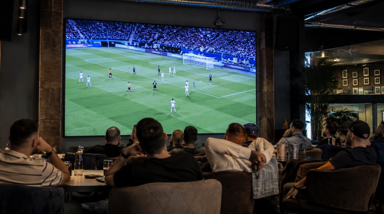 Grupa ljudi u modernom sportskom baru gleda fudbalsku utakmicu na velikom ekranu