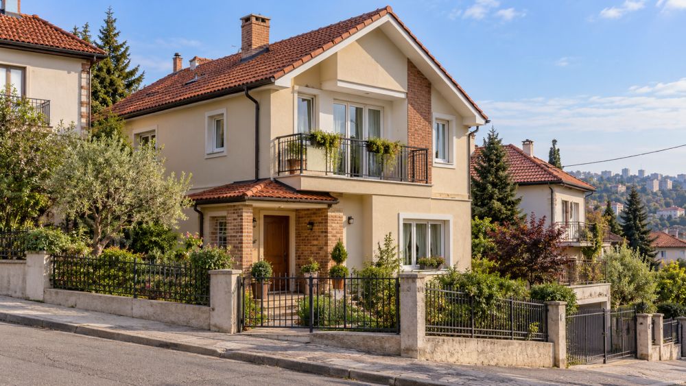 Porodična kuća na sprat sa bež fasadom, crvenim krovom, balkonom i ograđenim dvorištem u gradskom naselju.